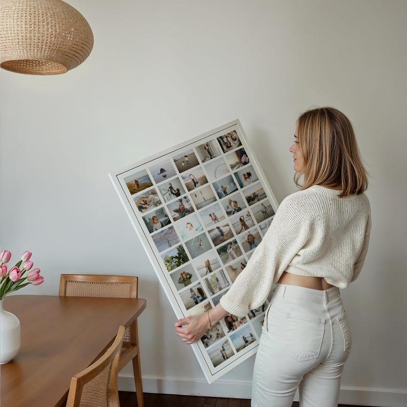 Op de afbeelding: Een grote, wit omkaderde fotocollage met talrijke kleine vierkante foto's. De collage wordt vastgehouden door een persoon in een cr&egrave;mekleurige trui en een witte broek. De achtergrond omvat een houten tafel, stoel en een geweven lamp.
