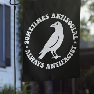 May include: A black rectangular garden flag featuring a white graphic of a crow in the center. The text "SOMETIMES ANTISOCIAL" is arched above the bird, and "ALWAYS ANTIFACIST" is arched below, with small star icons separating the phrases.