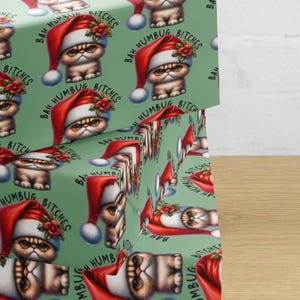 May include: Light green wrapping paper featuring a repeating pattern of a grumpy cat wearing a red and white Santa hat with a small holly sprig. The text "BAH HUMBUG BITCHES" is printed in black, arched over each cat illustration on the paper.