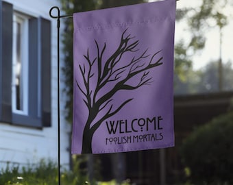 May include: A rectangular garden flag in a light purple hue featuring a black silhouette of a leafless tree on the left side. The text "WELCOME" is printed in a stylized black font above the words "FOOLISH MORTALS" in a matching black typeface.