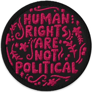 May include: A round, embroidered patch with the text "HUMAN RIGHTS ARE NOT POLITICAL" in pink. The text is surrounded by decorative floral elements, all on a black background. The patch has a black border.
