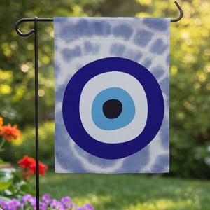 May include: A rectangular garden flag featuring a central evil eye symbol with concentric circles in dark blue, white, and light blue, set against a light blue and white tie-dye background, suspended from a black metal garden flag stand.