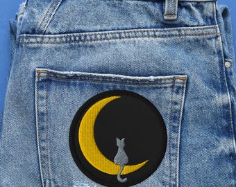 May include: Light blue denim jeans with a black and yellow embroidered patch on the back pocket. The patch features a black circle with a yellow crescent moon and a gray cat silhouette.