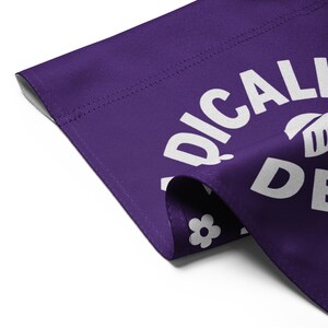 May include: A close-up of a purple fabric with white text that reads "RADICALLY DEEP". A white flower is visible in the corner. The fabric is folded over, showing the edge and stitching.