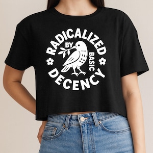 May include: A black short-sleeved crop top featuring a white circular graphic. The design includes a bird holding a branch in its beak, surrounded by the text "RADICALIZED BY BASIC DECENCY" and two small flower icons on the left and right sides.