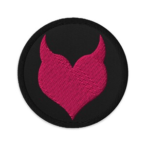 May include: A round, black embroidered patch featuring a pink heart-shaped design with two upward-curving horns. The heart and horns are filled with textured stitching, and the patch has a black border.