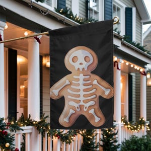 May include: A rectangular garden flag with a black background featuring a centered illustration of a light brown gingerbread person with a white skeletal bone design printed on its body, including a skull head and rib cage details.