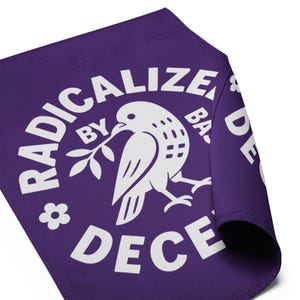 May include: A purple textile with a white graphic of a dove holding an olive branch, surrounded by the words "RADICALIZE BY DECENCY". The design is circular, with the text curving around the dove. A small flower graphic is also present.