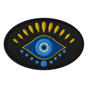 May include: Embroidered patch featuring a blue eye design on a black oval background. The eye has a blue iris with a white center, outlined in gold. Gold tear-shaped accents and blue dots surround the eye.