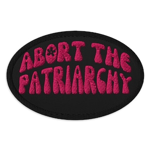 May include: An oval-shaped embroidered patch with the phrase "ABORT THE PATRIARCHY" in pink, stylized lettering. The patch has a black border and a small flower graphic. The patch is likely designed to be sewn or ironed onto clothing or accessories.