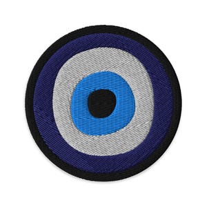May include: A circular embroidered patch featuring a concentric design of a black center, a bright blue ring, a white ring, a dark blue ring, and a thick black outer border. The embroidery texture is visible throughout the entire circular shape.