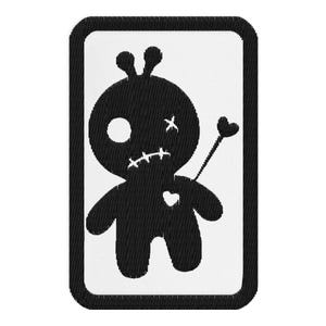 May include: A black and white embroidered patch featuring a voodoo doll design. The doll has a stitched mouth, a heart on its chest, and a heart-tipped arrow. The patch is rectangular with a black border.