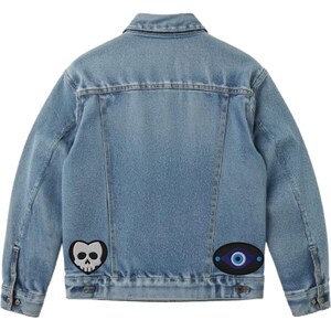 May include: Light blue denim jacket with two embroidered patches. One patch is a black heart with a white skull, and the other is an oval with a blue eye design. The jacket has a classic collar and button closures.