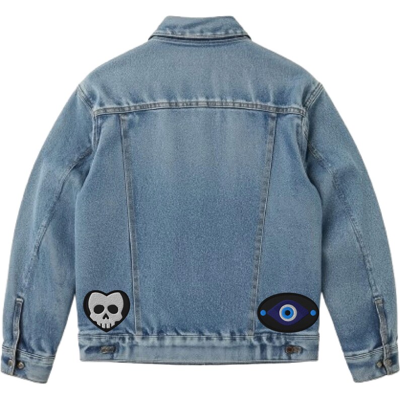 May include: Light blue denim jacket with two embroidered patches. One patch is a black heart with a white skull, and the other is an oval with a blue eye design. The jacket has a classic collar and button closures.