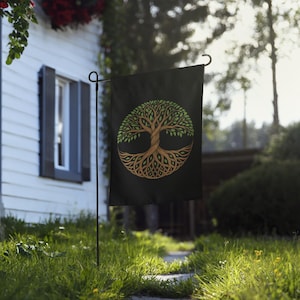 May include: A rectangular black garden flag featuring a circular graphic of a tree of life. The tree has a brown trunk and branches that intertwine with green leaves, forming a symmetrical pattern centered on the dark fabric of the flag.