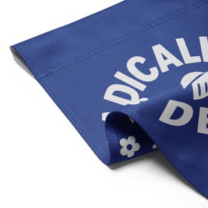 May include: A blue fabric item with white text that reads "RADICALLY DE". A white flower graphic is visible. The fabric is folded over, showing a seam and a white background.