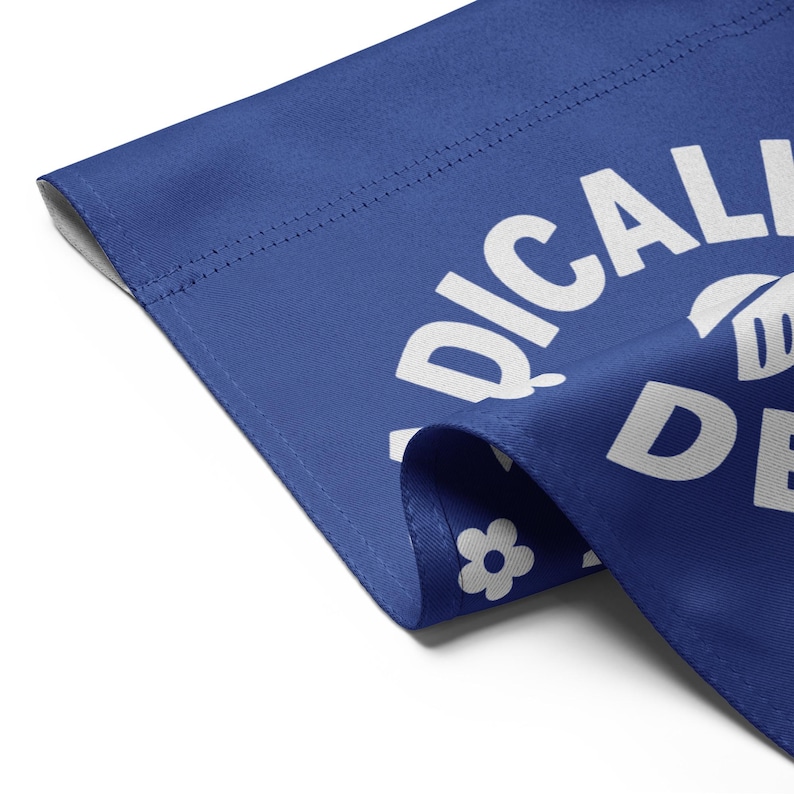 May include: A blue fabric item with white text that reads "RADICALLY DE". A white flower graphic is visible. The fabric is folded over, showing a seam and a white background.