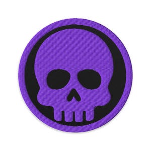 May include: A circular embroidered patch featuring a purple skull design within a black circle. The skull has black eye sockets and a jawline. The patch is on a white background.