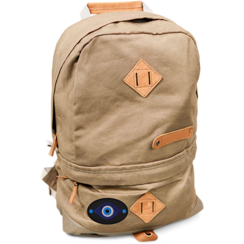 May include: A tan canvas backpack with leather accents. Features a diamond-shaped leather patch and a blue eye symbol. Includes a top handle and adjustable straps.