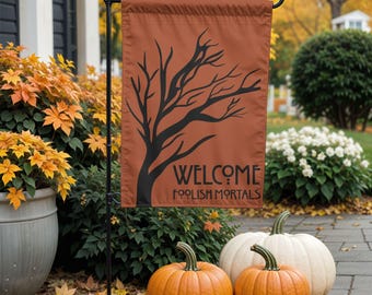 May include: A rectangular garden flag in a burnt orange color featuring a black silhouette of a bare tree and the text "WELCOME FOOLISH MORTALS" in a stylized black font, suspended from a black metal stand.