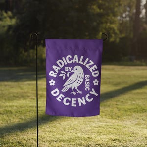 May include: A purple garden flag with the words "RADICALIZED BY BASIC DECENCY" in white, encircling a white dove holding an olive branch. The flag is displayed on a black metal stand in a grassy outdoor setting.
