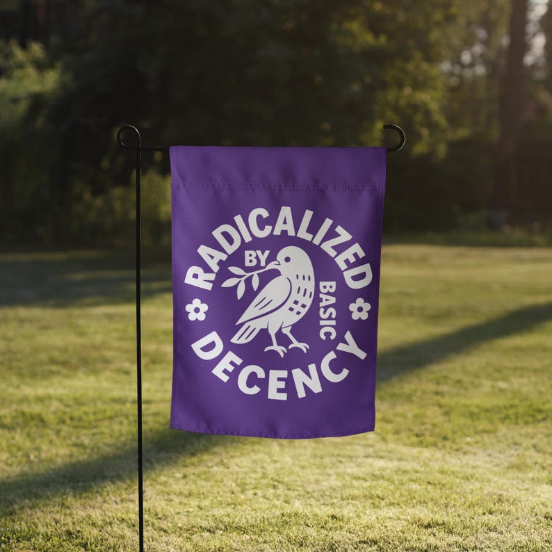 May include: A purple garden flag with the words "RADICALIZED BY BASIC DECENCY" in white, encircling a white dove holding an olive branch. The flag is displayed on a black metal stand in a grassy outdoor setting.