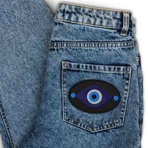 May include: Light blue denim jeans with a black patch on the back pocket. The patch features a blue eye design with a white and blue center, and two blue dots on either side. The jeans are folded.