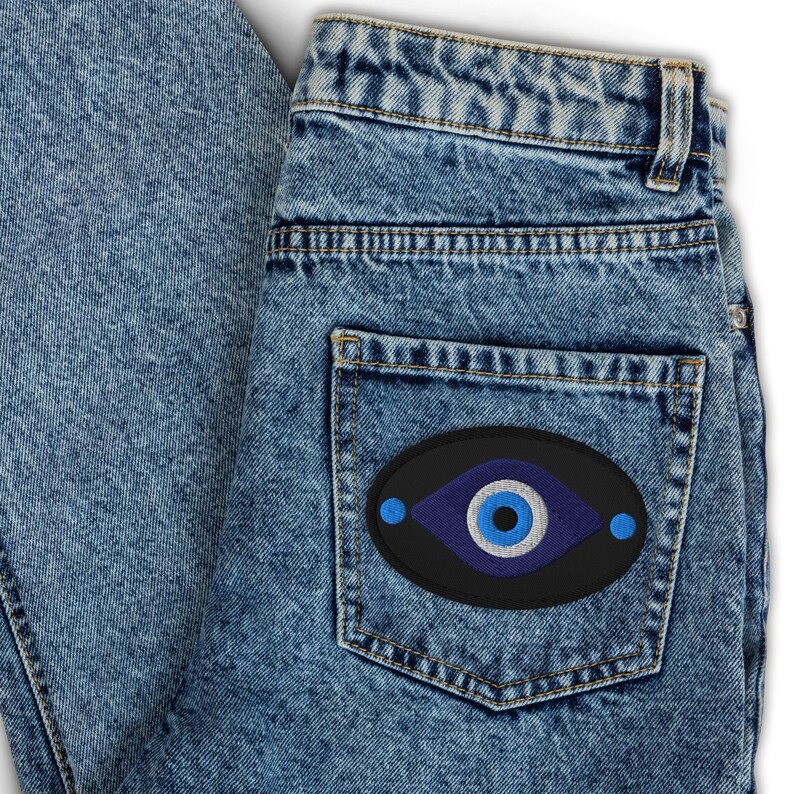 May include: Light blue denim jeans with a black patch on the back pocket. The patch features a blue eye design with a white and blue center, and two blue dots on either side. The jeans are folded.