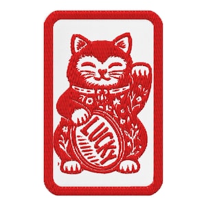 May include: Embroidered patch featuring a red Maneki-neko cat with a raised paw, a symbol of good luck. The cat holds an oval with the word "LUCKY" and is set against a white background with a red border. The design is detailed with floral patterns.