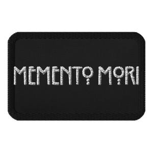May include: Black rectangular patch with the embroidered phrase "MEMENTO MORI" in white. The text is in a stylized font, and the patch has a stitched border.