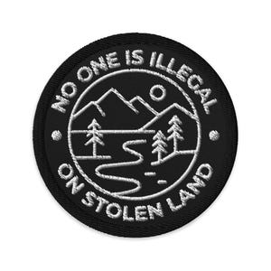 May include: Black embroidered patch with the text "NO ONE IS ILLEGAL ON STOLEN LAND" surrounding a white graphic of mountains, trees, a river, and a sun. The patch is circular and has a black border.