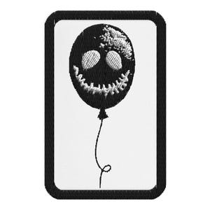May include: A black and white embroidered patch featuring a balloon with a spooky face design. The balloon has a stitched, toothy grin and eye details. The patch has a black border and a white background.