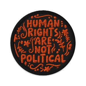 May include: Embroidered patch with the text "HUMAN RIGHTS ARE NOT POLITICAL" in orange thread on a black circular background. Decorative floral and leaf accents surround the text.
