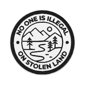May include: Embroidered patch with the text "NO ONE IS ILLEGAL ON STOLEN LAND" in a circular design. The patch features a mountain range, trees, a river, and a sun, all in black and white.