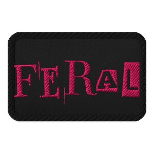 May include: Black embroidered patch with the word "FERAL" in bright pink, stylized lettering. The patch has a rectangular shape with rounded corners and a black border. The text is centered on the patch.