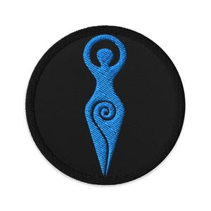 May include: A round, black embroidered patch featuring a blue stylized female figure with arms raised and a spiral design on the torso. The patch has a black border and is set against a white background.