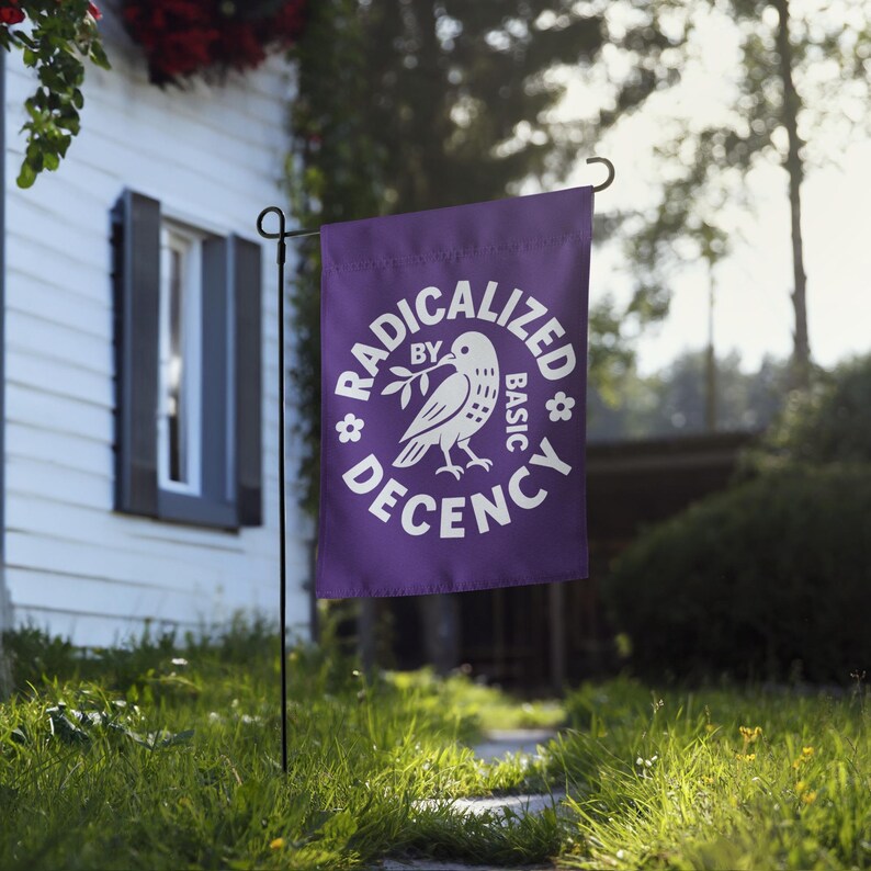 May include: A purple garden flag with the words "RADICALIZED BY BASIC DECENCY" in white, surrounding a white dove holding an olive branch. The flag is hanging on a black metal stand in a grassy yard.