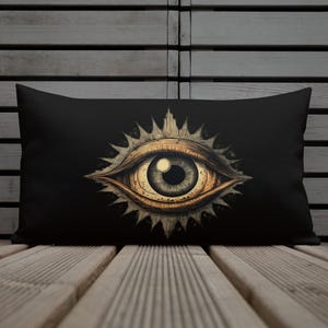 May include: Black rectangular pillow featuring a detailed illustration of an eye. The eye is in shades of brown and gold, with a sunburst design around it. The pillow is resting on a wooden surface.