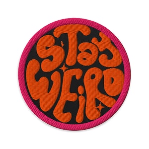 May include: Embroidered patch with the words "Stay Weird" in orange bubble letters on a black background. The patch has a pink border and small star details. This patch is perfect for jackets, bags, or other fabric items.