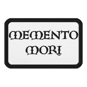 May include: A rectangular patch with a white background and a black border. The words "MEMENTO MORI" are embroidered in black, gothic-style lettering.