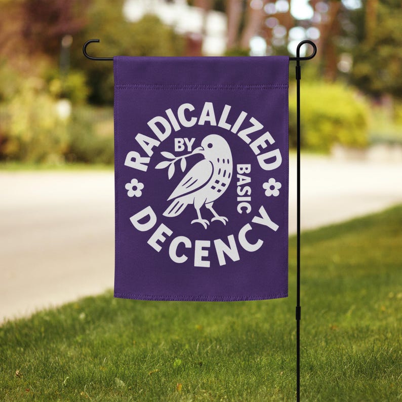 May include: A purple garden flag with the words "RADICALIZED BY BASIC DECENCY" in white, surrounding a white dove holding an olive branch. The flag is hanging on a black metal pole in a grassy yard.
