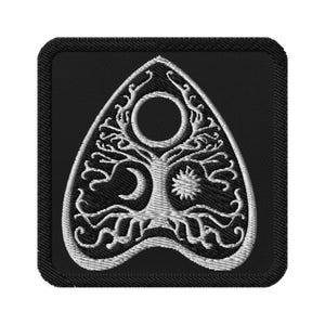 May include: Black embroidered patch featuring a white planchette design. The planchette has a tree of life design with a crescent moon, sun, and a central hole. The patch has a black border.