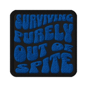 May include: Embroidered patch with the words "SURVIVING PURELY OUT OF SPITE" in blue, stylized lettering on a black background. The patch has a black border and a textured appearance.