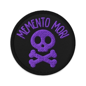 May include: Round black patch with purple embroidered skull and crossbones. The words "MEMENTO MORI" are embroidered above the skull. The patch has a black border.
