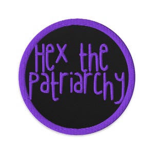 May include: A round embroidered patch with a black center and a purple border. The phrase "Hex the Patriarchy" is embroidered in purple, stylized lettering across the center of the patch.