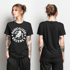 May include: Black t-shirt with a white graphic design. The design features a bird holding a branch, surrounded by the words "RADICALIZED BY BASIC DECENCY". The shirt is a crew neck style.