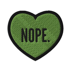 May include: Embroidered heart-shaped patch with a green textured fill and a black border. The word "NOPE." is embroidered in bold black letters across the center of the heart. The patch is designed to be sewn or ironed onto fabric.