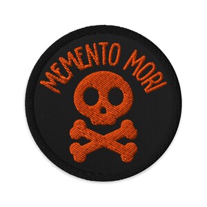 May include: Round black patch with orange embroidered skull and crossbones. The words "MEMENTO MORI" are embroidered in orange around the skull. The patch has a black border.