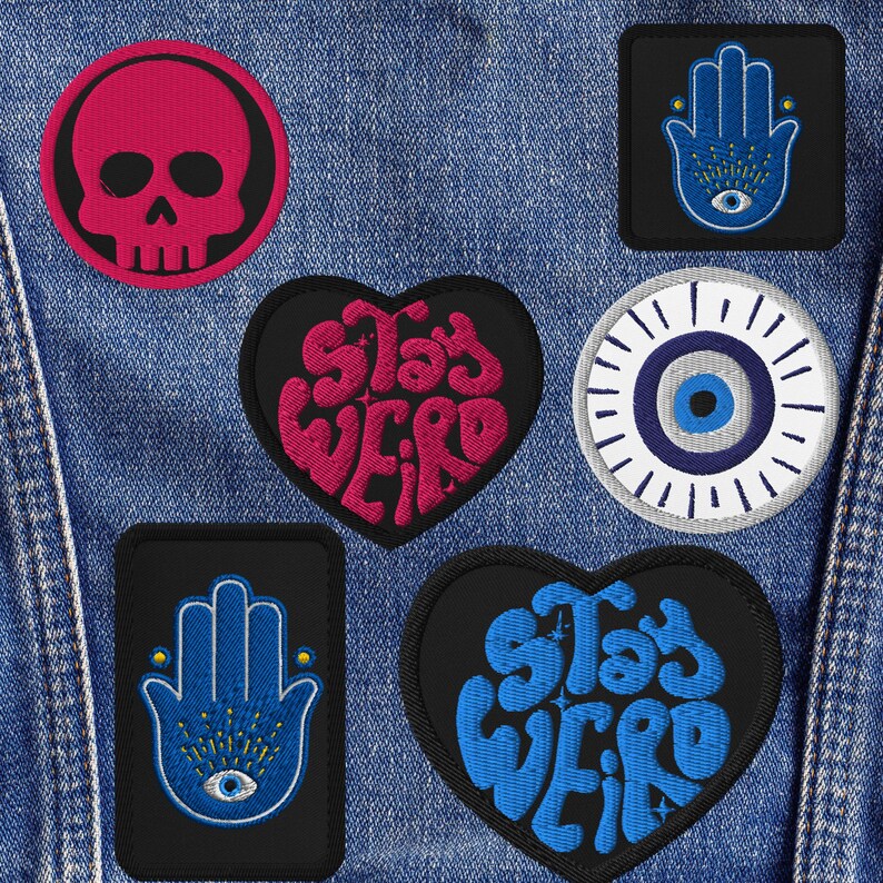 May include: A collection of embroidered patches in various shapes, including a skull, hearts with "Stay Weird" text, an eye, and Hamsa hand designs. The patches feature black, pink, and blue colors, with intricate stitching details. They are displayed on a denim background.
