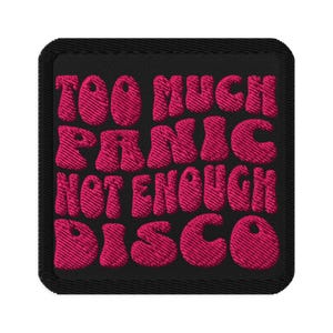 May include: A square, black embroidered patch with the phrase "TOO MUCH PANIC NOT ENOUGH DISCO" in pink, bubble-style lettering. The patch has a black border and a textured appearance.
